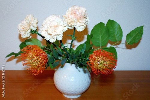 Wunderschöner Blumenstrauß aus Nadelkissen (Leucospermum) und Nelken (Dianthus) mit orangefarbenen und blassrosa Blüten in einer weißen Blumenvase. Die Tischplatte besteht aus Mahagoniholz. Nahaufnahm photo