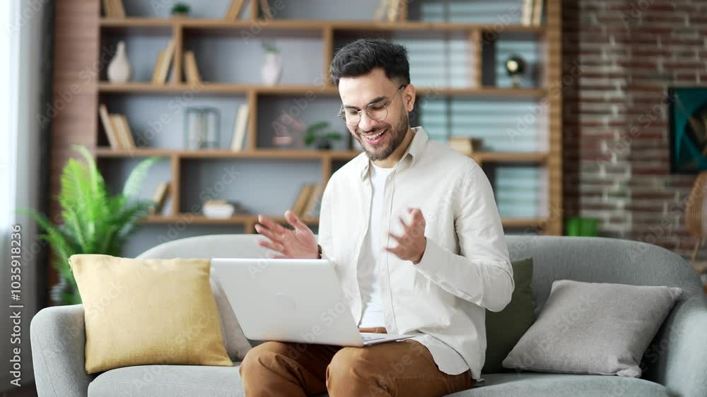 Young adult man talking on a video using a laptop sitting on sofa in living room in home office. Freelancer has a business meeting. A smiling coach speaks remotely at an online course or training Young adult man talking on a video using a laptop sitting on sofa in living room in home office. Freelancer has a business meeting. A smiling coach speaks remotely at an online course or training
