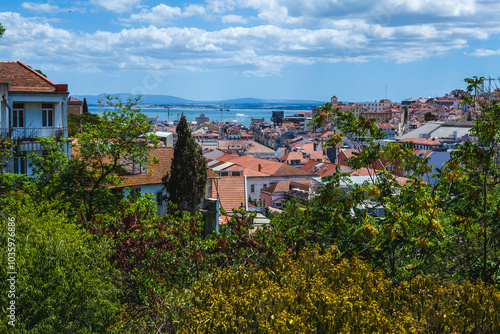 a scenic view overlooking a cityscape with red-tiled rooftops and traditional architecture, extending toward a large body of water in the background