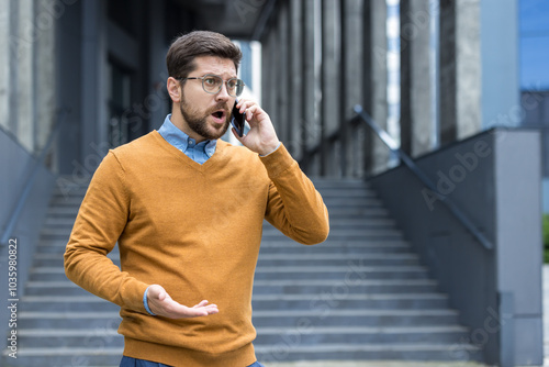 Upset young male businessman standing outside an office building, talking on the phone emotionally and angrily