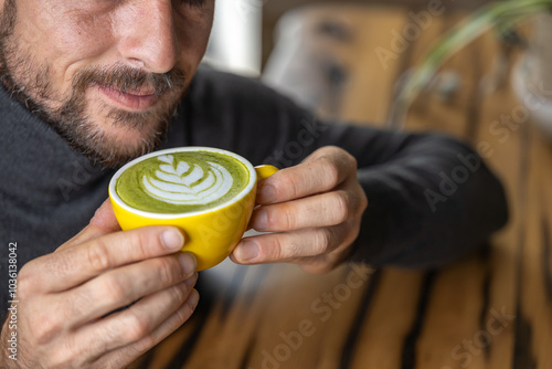 Close-up happy young man smiling in a cafe holding a cup of matcha tea with raspberry, heart-shaped latte art, cappuccino coffee with red foam and heart, decoration with plants, vegan hot drink