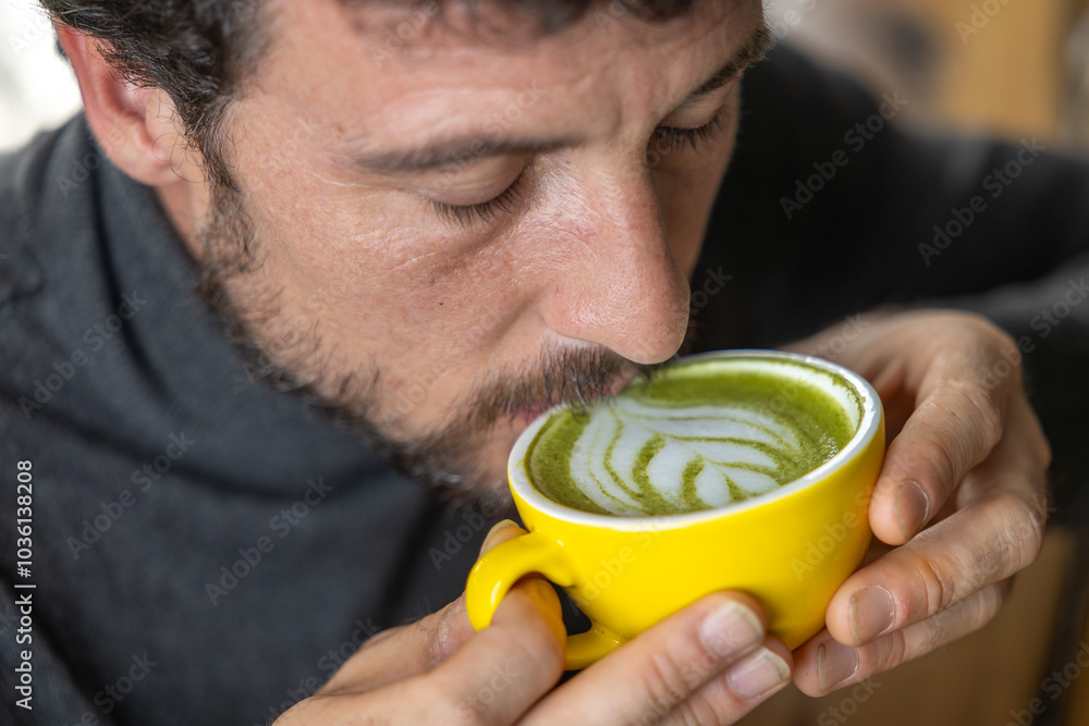 Close-up happy young man smiling in a cafe holding a cup of matcha tea with raspberry, heart-shaped latte art, cappuccino coffee with red foam and heart, decoration with plants, vegan hot drink Close-up happy young man smiling in a cafe holding a cup of matcha tea with raspberry, heart-shaped latte art, cappuccino coffee with red foam and heart, decoration with plants, vegan hot drink