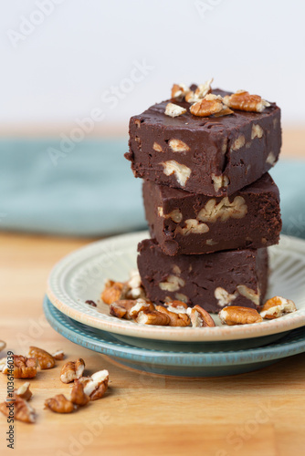 Stack of Three Pieces of Homemade Chocolate Pecan Fudge on a Plate with Pecan Pieces Scattered Around on a Kitchen Countertop