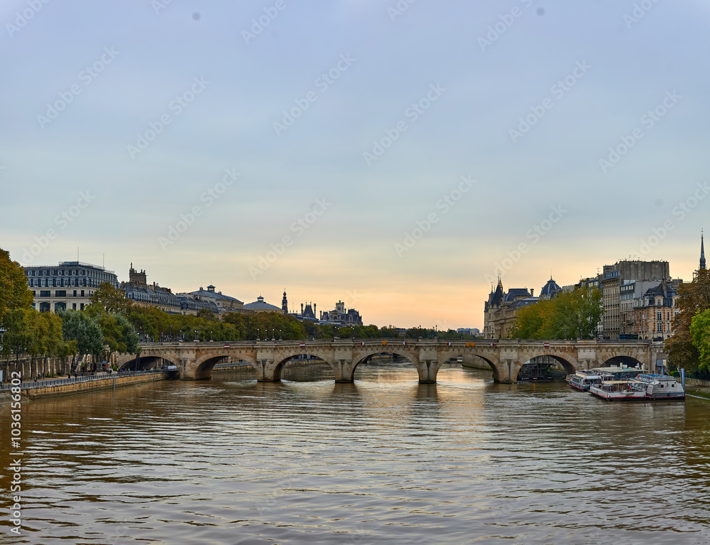 Fototapeta premium Seine River in Paris with stone bridge spanning the water, lined with historic buildings and trees, boats moored along the quay, overcast sky, calm water