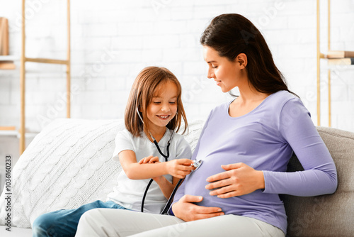 Little pretty girl listening belly of her pregnant mother through stethoscope, playing doctor at home, free space. Family and new life concept.