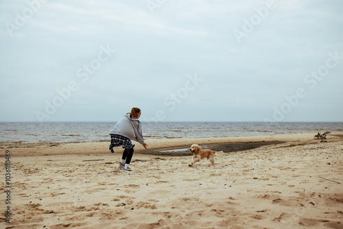 Obraz na płótnie Girl and dog playing by the seaside. The dog happily returns with the ball, capturing the playful bond between them in a serene autumn beach setting. w salonie