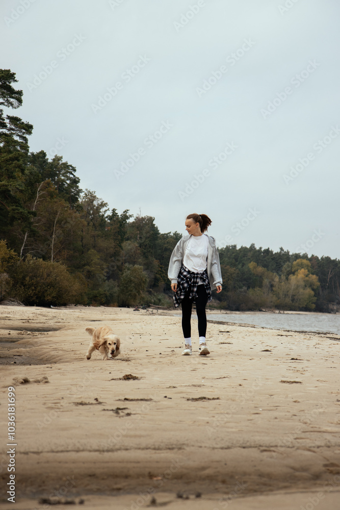 A girl in a gray hoodie and plaid shirt tied around her waist strolls along an empty beach with her light-colored Cocker Spaniel, enjoying the autumn breeze and the sound of the waves. Vertical photo