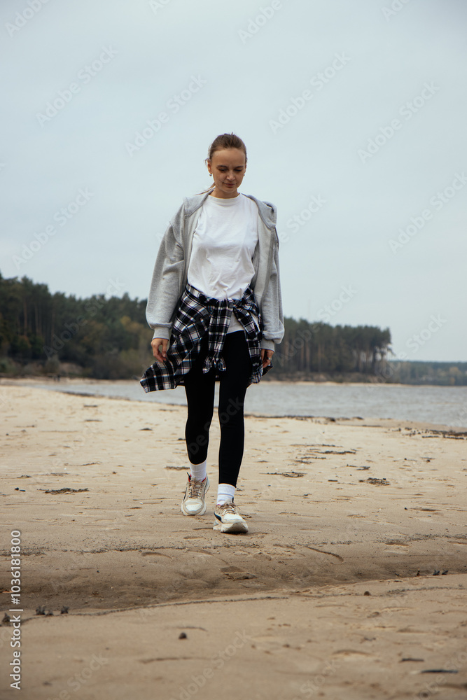 A girl in a gray sweatshirt and a black-and-white plaid shirt tied around her waist strolls along the beach, with the ocean waves and lush forest creating a picturesque backdrop. Vertical photo