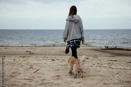 Woman enjoying a beach walk with her dog, dressed in cozy autumn clothes. Plaid shirt around her waist adds a relaxed, casual vibe. Ocean shore in the background.