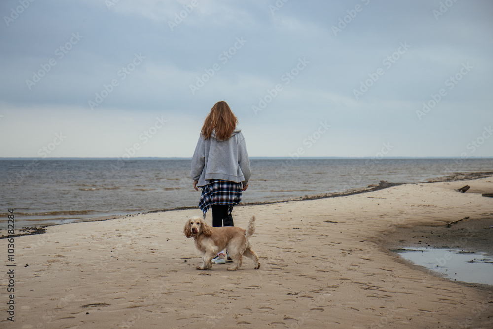 Girl walking her dog along the beach on a cloudy day. Casual autumn outfit with a plaid shirt tied around her waist. Outdoor recreation with a pet by the sea.