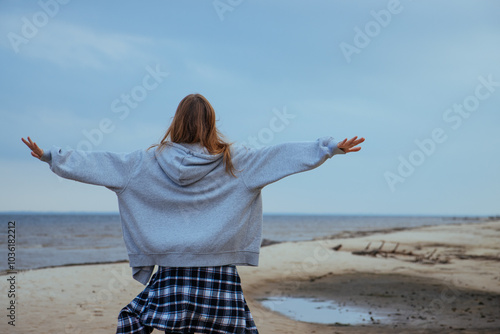 Obraz na płótnie Young woman walking on a sandy beach with her arms outstretched, embracing the feeling of freedom and nature. Serene autumn day by the sea, symbolizing peace and liberation. w salonie