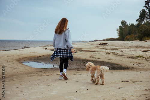 Woman walking her dog along the shore, wrapped in warm autumn clothing. The serene beach setting creates a peaceful outdoor mood, perfect for themes of nature and pet care.