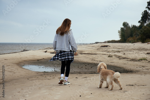 Obraz na płótnie Woman walking her dog along the shore, wrapped in warm autumn clothing. The serene beach setting creates a peaceful outdoor mood, perfect for themes of nature and pet care. w salonie