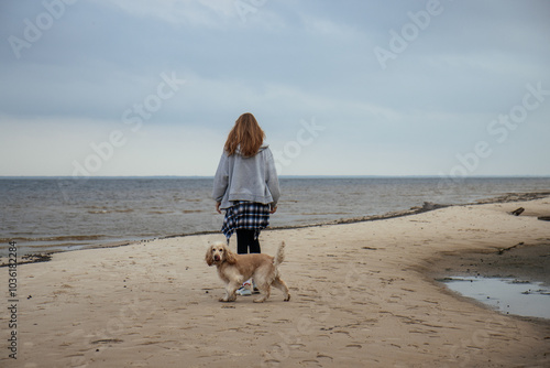 Obraz na płótnie Girl walking her dog along the beach on a cloudy day. Casual autumn outfit with a plaid shirt tied around her waist. Outdoor recreation with a pet by the sea. w salonie