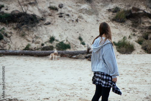 Obraz na płótnie Girl looks on as her dog explores the terrain by the beach. The dog stands on a log, surrounded by sand and plants, showcasing a calm autumn day in nature. w salonie