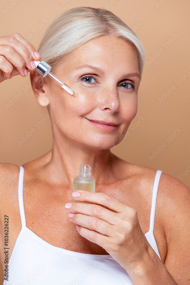 Skincare beauty portrait of a mature senior woman with a grey silver hair and glowing skin applying a facial care serum or retinol with a pipette dropper against beige background. Skincare beauty portrait of a mature senior woman with a grey silver hair and glowing skin applying a facial care serum or retinol with a pipette dropper against beige background.