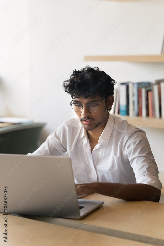 Focused busy young Indian business professional man working at laptop in office, sitting at table, typing. Serious smart male student using Internet technology in library. Vertical portrait Focused busy young Indian business professional man working at laptop in office, sitting at table, typing. Serious smart male student using Internet technology in library. Vertical portrait
