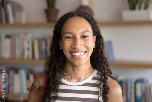 Cheerful beautiful teenager African American student girl posing for head shot photo in public library, standing with bookshelves in background, looking at camera with toothy smile