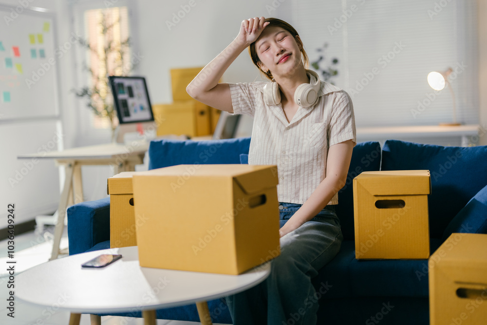 Young woman taking a break and wiping sweat from her forehead while sitting on a blue sofa surrounded by cardboard boxes Young woman taking a break and wiping sweat from her forehead while sitting on a blue sofa surrounded by cardboard boxes