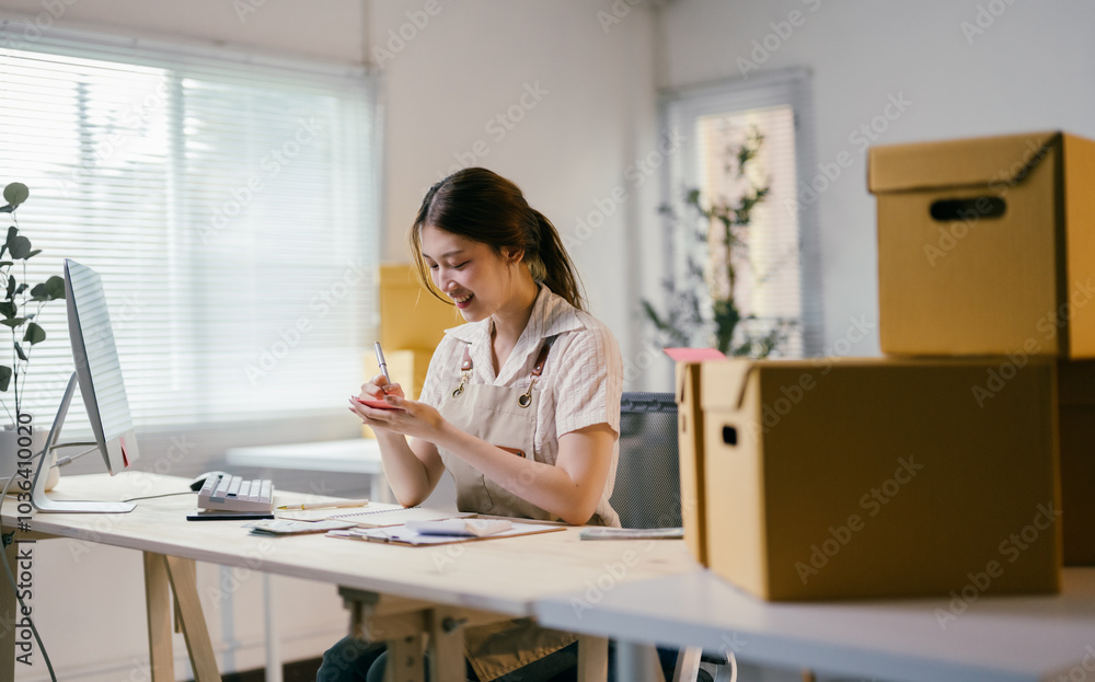 Young asian entrepreneur writing down notes while managing her business from home office, surrounded by cardboard boxes Young asian entrepreneur writing down notes while managing her business from home office, surrounded by cardboard boxes