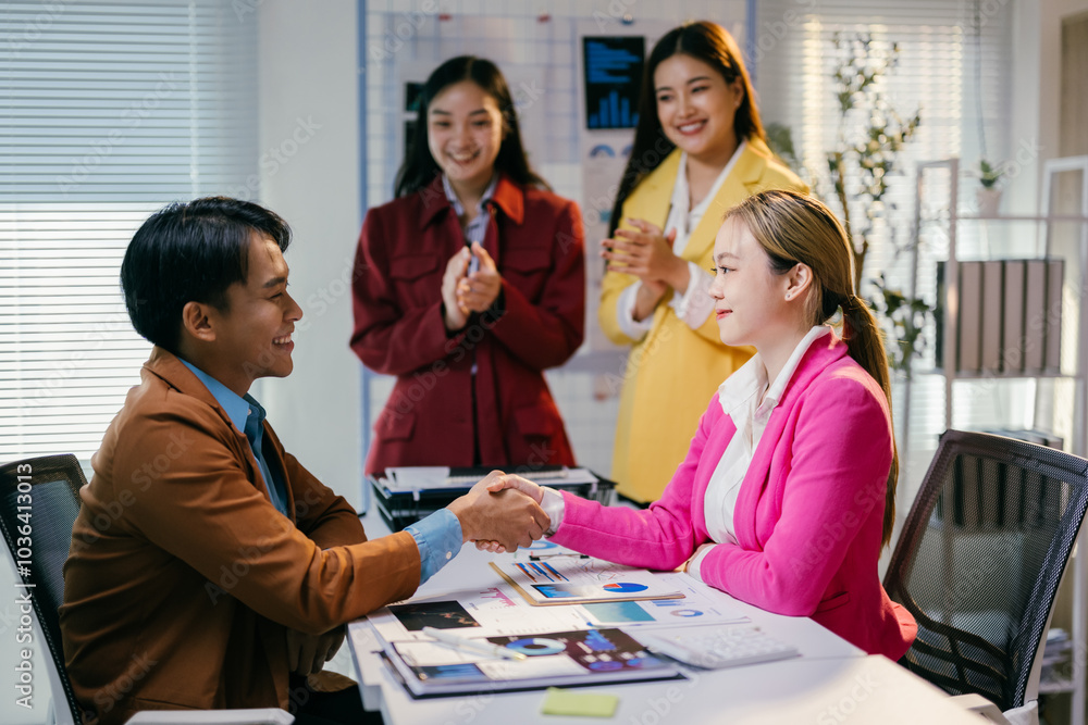 Young businessman and businesswoman shaking hands after closing a deal in a modern office, their teammates are applauding them in the background Young businessman and businesswoman shaking hands after closing a deal in a modern office, their teammates are applauding them in the background