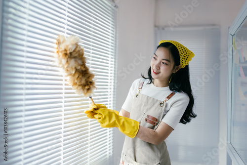 Cheerful woman cleans blinds with a feather duster, wearing yellow gloves and smiling, bringing freshness to her home photo