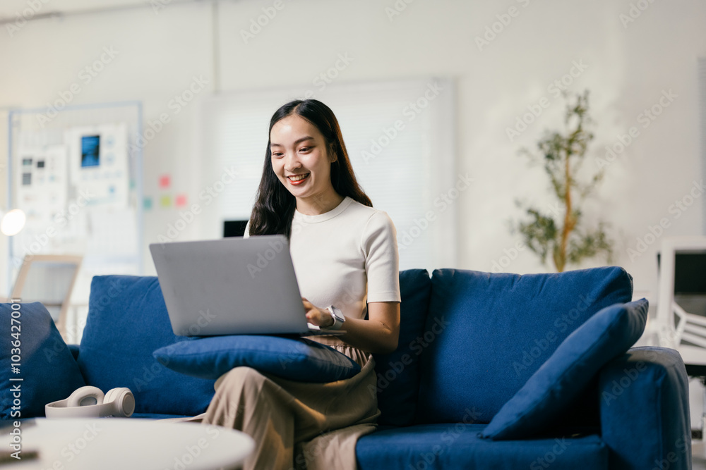 Smiling young woman is working remotely from her home office, comfortably sitting on a blue sofa while typing on her laptop Smiling young woman is working remotely from her home office, comfortably sitting on a blue sofa while typing on her laptop
