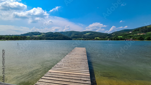 Wooden pier extends into the calm waters of the lake Stubenbergsee surrounded by rolling hills of East Styrian Hill Country, Styria, Austria. Lush green forest on foothills of Prealps East of the Mur photo