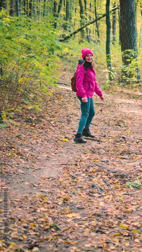 Attractive young woman in a pink jacket, jeans, and brown backpack walking through an autumn forest, turns, smiles at camera and gestures follow me or let's go with her hand, slow motion Attractive young woman in a pink jacket, jeans, and brown backpack walking through an autumn forest, turns, smiles at camera and gestures follow me or let's go with her hand, slow motion