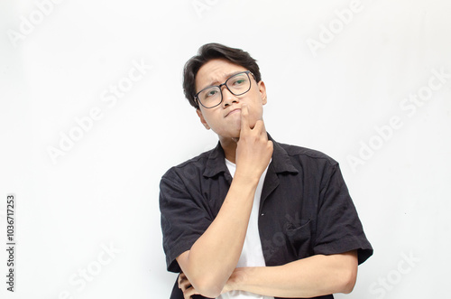 Thoughtful asian man thinking and wondering with hand touching skin as thinking gesture isolated over white background. asian man with glasses and black and white casual shirt feeling uncertain. photo