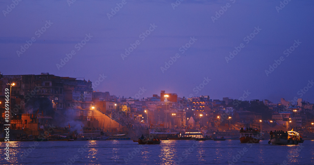 Varanasi, Uttar Pradesh, India. Funeral rites of cremation at the Harishchandra Ghat. Evening pink purple sunset sky, night time. Camera moving along riverbank embankment. Cinematic View from boat