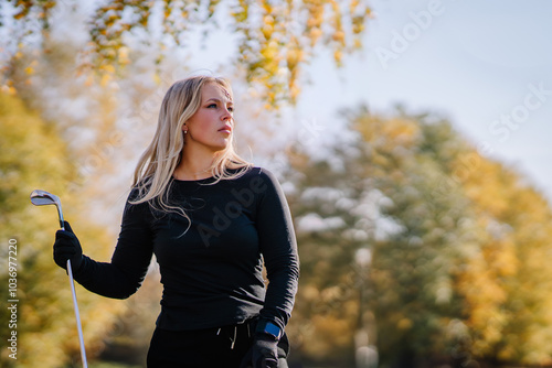 A woman with fake blood on her face, holding a golf club, standing outdoors on a sunny day with autumn trees in the background. Halloween theme.. photo