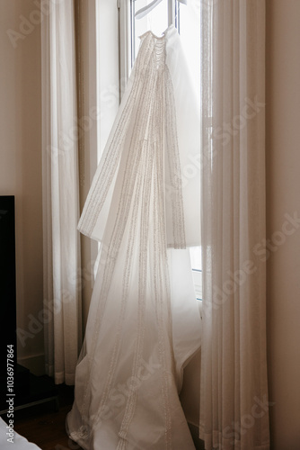 Elegant white wedding dress with long veil hanging by a cozy window in a minimalist bedroom setting.