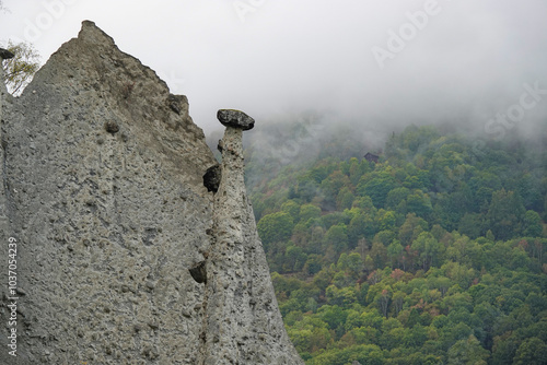 CLOSE UP: Fairy chimney of Les Pyramides dEuseigne rises dramatically from the mist. Unique and naturally eroded rock formation standing tall above lush green alpine forest partially shrouded by fog. photo