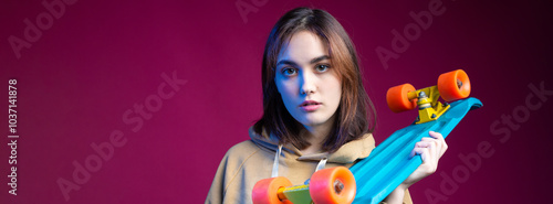 Young beautiful fashionable hipster girl dressed in a hoodie holds a skateboard in her hands on a purple studio background.