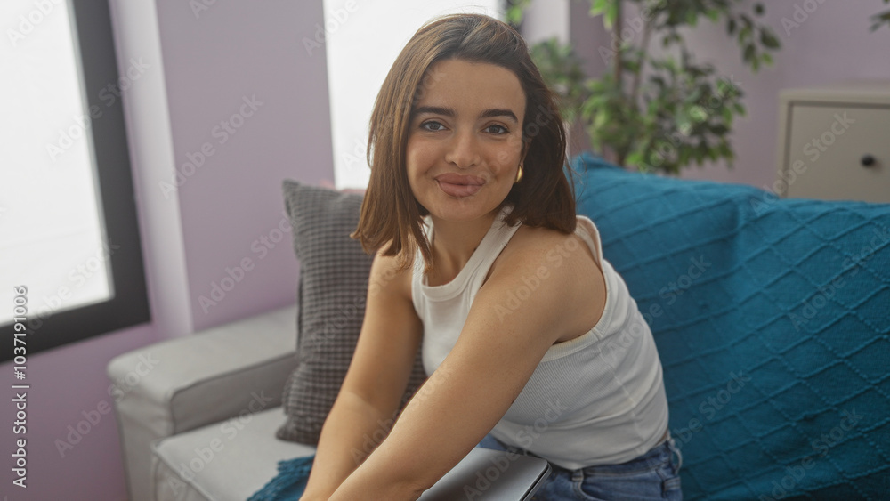 Beautiful hispanic brunette woman sitting on a sofa in a cozy living room with purple walls and blue pillows indoors, smiling warmly at the camera Beautiful hispanic brunette woman sitting on a sofa in a cozy living room with purple walls and blue pillows indoors, smiling warmly at the camera