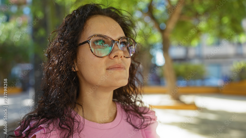 Young woman in glasses standing outdoors in urban park with green trees and blurred city background, looking thoughtful and content. Young woman in glasses standing outdoors in urban park with green trees and blurred city background, looking thoughtful and content.