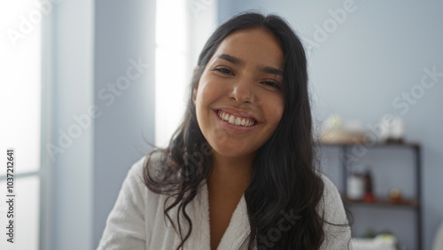 Young woman smiling in a wellness center wearing a robe, exuding beauty and calm in an indoor spa setting with soft lighting and minimalistic decor with shelves in the background.