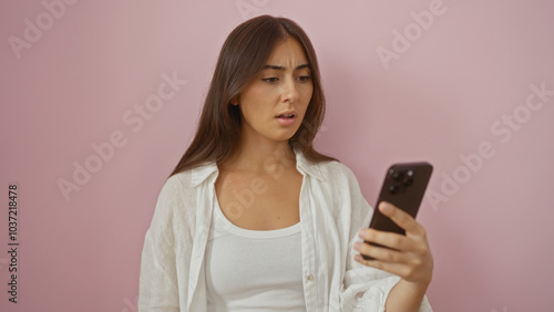 Young attractive hispanic brunette woman over isolated pink background looking at smartphone with a concerned expression