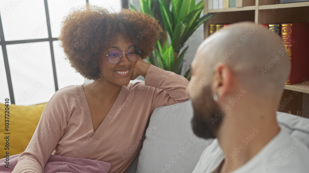 Fototapeta premium Interracial couple smiling lovingly at each other indoors, with a woman and a bald man on a couch, suggesting a comfortable, affectionate relationship.