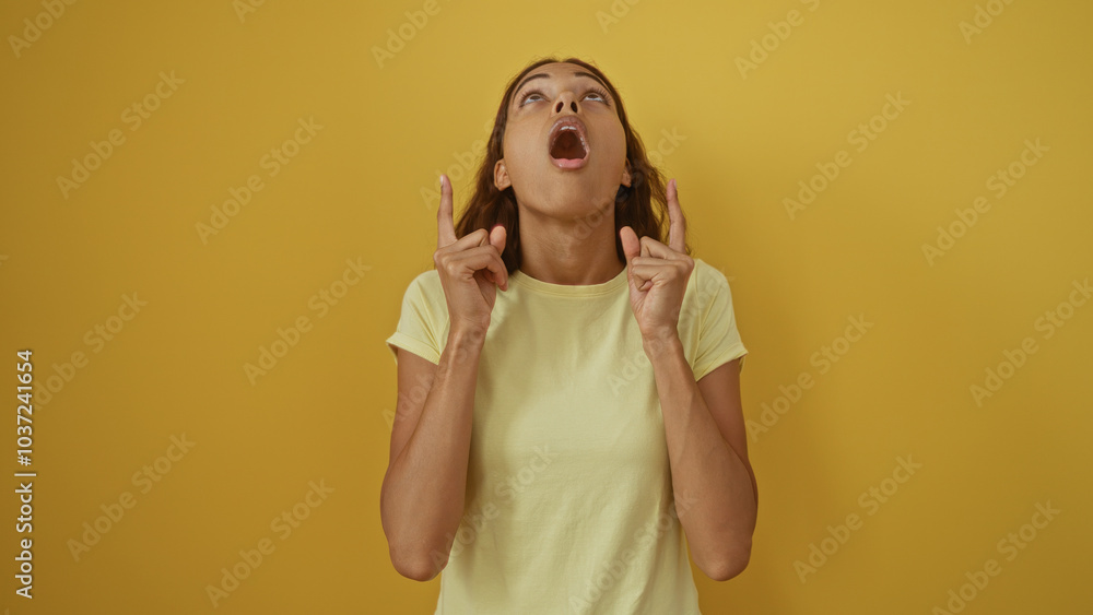 Fototapeta premium Young woman with brunette hair looking upwards in surprise, pointing with fingers, over an isolated yellow wall background.