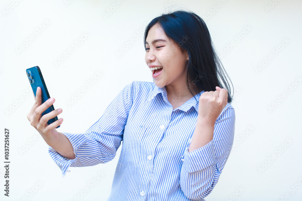 happy and excited asian woman holding smartphone celebrate winning victory by raising fist wearing striped shirt isolated on white background happy and excited asian woman holding smartphone celebrate winning victory by raising fist wearing striped shirt isolated on white background