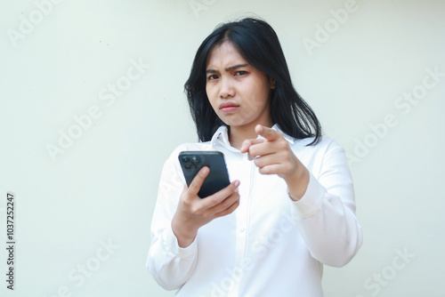 portrait of sad unbelieve asian business woman pointing to camera with glare eye holding smartphone, disappointment and disatisfied expression, wear formal white suit standing over isolated background photo