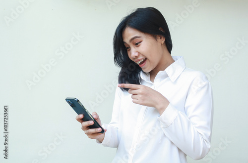 successful playful asian young saleswoman surprised looking to smartphone hold on hands wearing white formal suit shirt, pointing index finger to screen, standing isolated