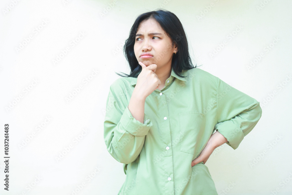 thoughtful asian woman with dark hair looking away suspicious with hands on chin and waist wearing green oversized shirt isolated on white background thoughtful asian woman with dark hair looking away suspicious with hands on chin and waist wearing green oversized shirt isolated on white background