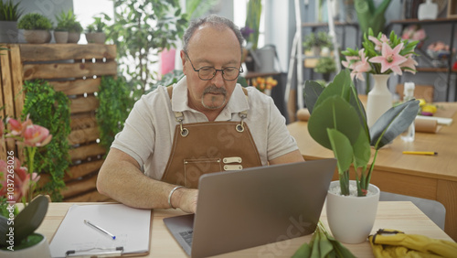 A mature man works on a laptop in a flower shop surrounded by plants and flower arrangements.