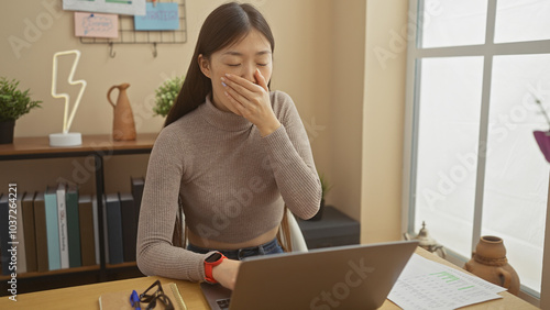 Tired asian woman yawning while working on laptop in home office setting