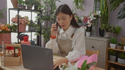 Obraz na plátně A chinese woman florist in an apron uses a laptop and phone inside a flower shop surrounded by fresh bouquets and plants