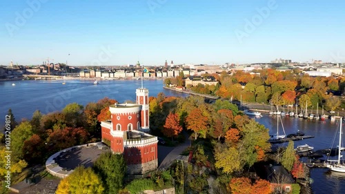 Aerial arc view of Kastellholmen red castle on island with Stockholm skyline in the background on autumn morning with turning yellow and orange trees in Sweden and Swedish flag waving in the wind photo