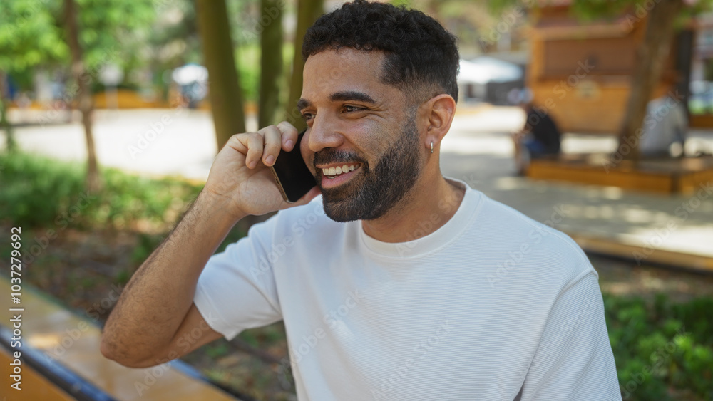 Young man talking on smartphone in urban park, smiling under the shade of trees with a blurred city background Young man talking on smartphone in urban park, smiling under the shade of trees with a blurred city background